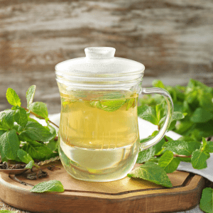  A glass cup of freshly brewed lemon balm tea with a lid on top, surrounded by fresh lemon balm leaves on a wooden tray against a rustic background.